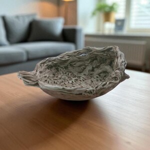 Op de afbeelding: Een decoratieve, getextureerde kom met een natuurlijke, organische uitstraling. De kom heeft een gedempte grijsgroene kleur en staat op een lichtbruine houten tafel. Het ontwerp van de kom heeft een gedetailleerd, open patroon.