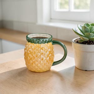 Może przedstawiać: Żółty ceramiczny kubek w kształcie ananasa o teksturowanej powierzchni w romby. Kubek posiada ciemnozielony brzeg oraz pasujący ciemnozielony uchwyt. Wnętrze kubka jest białe i wypełnione ciemnym napojem w płynnej postaci.