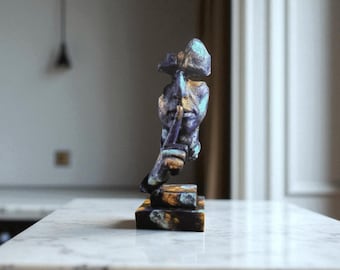 Könnte beinhalten: Eine bronzefarbene Skulptur einer Figur mit einer Hand am Mund, die Stille andeutet. Die Statue hat eine strukturierte Oberfläche mit blauen und goldenen Akzenten. Sie steht auf einem rechteckigen Sockel mit ähnlichen Farbvariationen.