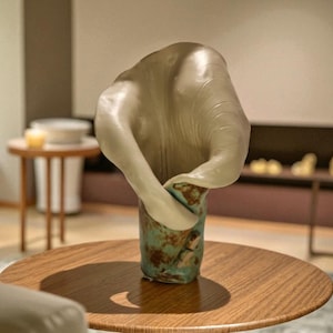 Puede incluir: Un jarrón de cerámica abstracto con una parte superior crema en forma de pétalo y una base turquesa y marrón. El jarrón se exhibe sobre una mesa redonda de madera, mostrando su diseño y textura artísticos. Un artículo de decoración del hogar.