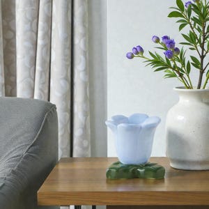 May include: A wooden table displays a light blue tulip-shaped glass, a white speckled vase with purple flowers, and a green leaf-shaped glass base. The background features a gray armchair and patterned curtains.