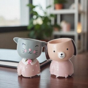 Può includere: Due fioriere in ceramica a forma di animali dei cartoni animati. Uno è un gatto grigio con dettagli rosa e un vestito rosa. L'altro è un cane marrone con orecchie marroni e un vestito rosa.