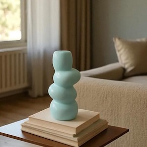 Może przedstawiać: Jasnoniebieski wazon ceramiczny o abstrakcyjnym kształcie. Wazon stoi na stosie książek na brązowym stoliku. Tło obejmuje okno z zasłonami i beżową sofę.