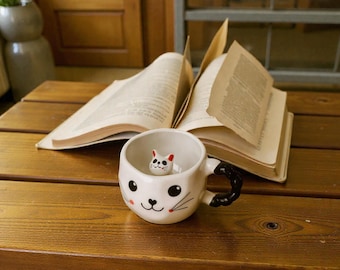 Puede incluir: Una taza de cerámica blanca con temática de gato, con asa negra y una pequeña figura de gato en el interior. La taza está colocada sobre una superficie de madera con un libro abierto en el fondo. La taza tiene bigotes negros y una cara sonriente.