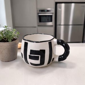 May include: A white ceramic mug with a black geometric pattern and a black handle. The mug is on a white surface beside a small potted plant with green leaves. The background shows a kitchen with stainless steel appliances.