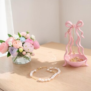 May include: A display of a floral arrangement in a glass vase, a pink bow-shaped jewellery holder, and a gold and pearl bracelet. The flowers are pink, purple, and white. The items are on a wooden surface.