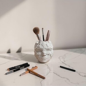 May include: A white, sculpted face-shaped container holding makeup brushes. Several makeup pencils and brushes are scattered on the white marble surface. The container and makeup items are lit by natural light, creating shadows.