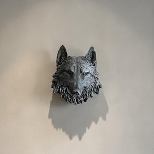 Puede incluir: Escultura detallada de una cabeza de lobo gris montada en una pared. La escultura tiene una superficie texturizada, que captura el pelaje y los rasgos faciales del lobo. La pared es de color beige neutro y la escultura proyecta una sombra.