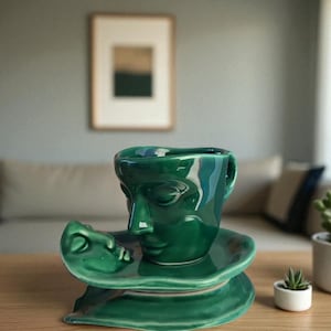 Può includere: Un set di tazza e piattino da tè in ceramica verde con un design a volto. La tazza da tè presenta un volto scolpito e il piattino ha un design a volto abbinato. Il set è posto su un tavolo di legno con una piccola pianta succulenta.