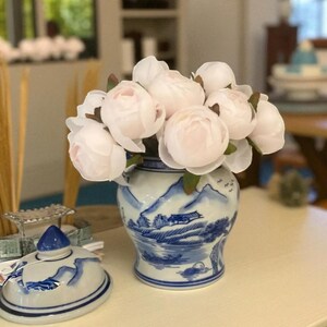Puede incluir: Un jarrón de cerámica blanco y azul con un diseño de paisaje, lleno de peonías de color rosa claro. Un recipiente con tapa a juego está al lado del jarrón. En el fondo hay hierbas secas decorativas y otros objetos.