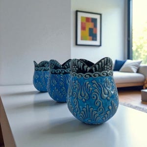 Könnte beinhalten: Drei blaue Keramikvasen mit Tulpenform und aufwendigen, geprägten floralen Designs. Die Vasen haben einen gewellten Rand mit einem dunkelgrauen Rand. Die Vasen werden auf einer weißen Oberfläche ausgestellt.