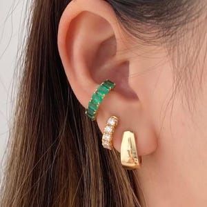 Peut inclure: Gros plan sur trois bijoux : une manchette d'oreille dorée avec des pierres vertes, une petite boucle d'oreille créole dorée et une boucle d'oreille dorée avec des pierres transparentes. Les bijoux sont portés sur l'oreille d'une personne aux longs cheveux bruns.