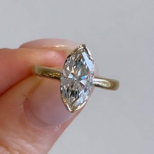 May include: A marquise-cut diamond ring with a gold band. The diamond is set in a bezel setting, and the band is thin and polished. The ring is held up against a plain background.