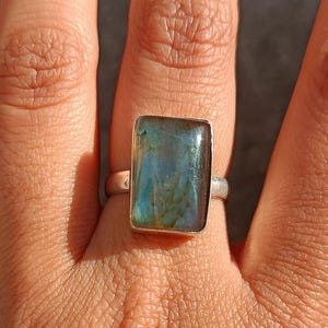 Puede incluir: Un anillo de plata con una piedra preciosa rectangular iridiscente azul-verdosa. El anillo se muestra en una mano, destacando las variaciones de color de la piedra. La banda del anillo es simple y delgada, resaltando el tamaño y la forma de la gema.