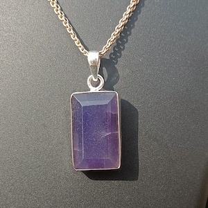 May include: A silver-toned chain necklace featuring a rectangular, faceted purple gemstone pendant set in a simple metal frame. The pendant hangs from a delicate link chain, showcasing a deep violet hue with subtle internal texture and light reflection.