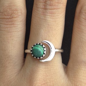 Peut inclure: Bague en argent ornée d'un croissant de lune et d'une pierre ronde en malachite verte. La bague est mise en valeur sur un fond sombre, soulignant les détails du bijou.
