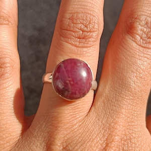 Peut inclure: Une bague en argent ornée d'une grosse pierre précieuse ronde et rouge foncé. La bague est portée sur une main à la peau claire, les doigts légèrement courbés. La pierre précieuse a une surface polie et lisse. La bague est de conception simple et élégante.