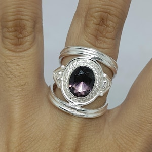 Puede incluir: Un anillo de plata con una piedra preciosa ovalada de color púrpura oscuro facetada, engastada en un borde de estilo cuerda trenzada. La banda consta de dos aros de plata separados que se unen en el engaste, creando un espacio abierto arriba y abajo.