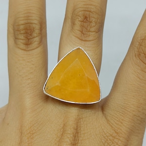 May include: A ring featuring a large, triangular-shaped, faceted orange gemstone set in a simple silver-toned metal band. The stone has a smooth, polished surface with subtle variations in its warm, vibrant orange hue and a clean, geometric cut.