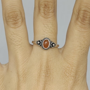 May include: A silver ring featuring a small, oval-shaped, reddish-brown stone set in the centre. The metal band displays a twisted rope texture around the stone setting, complemented by decorative silver bead accents on both sides of the centre piece.