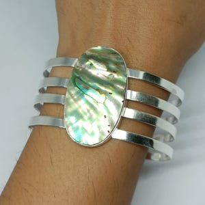 Puede incluir: Una pulsera tipo brazalete de tono plateado con una gran incrustación ovalada de concha de abulón. La pulsera está construida con cuatro bandas metálicas paralelas que se conectan a la pieza central iridiscente, mostrando tonos verdes, azules y nacarados.