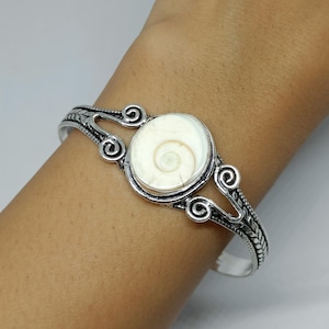 May include: A silver cuff bracelet featuring a central circular white shiva eye shell inlay. The metal band displays intricate oxidized scrollwork and filigree detailing on both sides of the stone, creating a textured and ornate appearance.