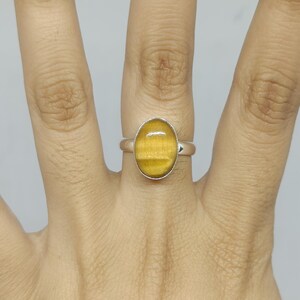 May include: A silver ring featuring a smooth, oval-shaped golden-brown tiger's eye gemstone set in a simple metal bezel. The stone displays a characteristic chatoyant luster with horizontal bands of light and dark amber tones across the surface.