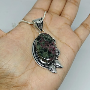 May include: An oval-shaped ruby zoisite gemstone pendant set in a polished sterling silver frame. The setting features decorative metal leaf accents and a textured bail, suspended from a matching silver chain necklace with a smooth finish.