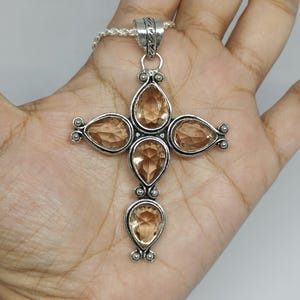 Puede incluir: Un colgante de cruz de plata con cuatro piedras preciosas de color marrón claro, talladas en forma de pera y dispuestas en cruz. El engaste de metal tiene un acabado texturizado y oxidado con pequeñas cuentas decorativas de plata.