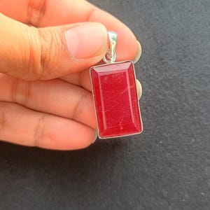 Puede incluir: Un colgante rectangular de piedra preciosa roja con un marco de metal plateado. El colgante se sostiene en una mano sobre un fondo oscuro. La piedra preciosa tiene una superficie pulida y un color rojo vibrante.