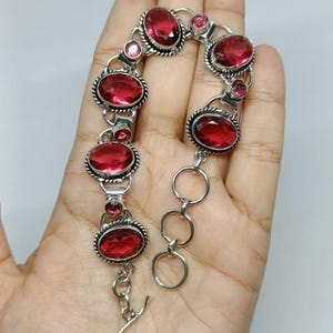 May include: A silver-toned bracelet featuring five large oval-cut deep red gemstones and four smaller round-cut red stones. The links are connected by twisted metal borders, and the piece includes a circular link extension chain with a toggle clasp.