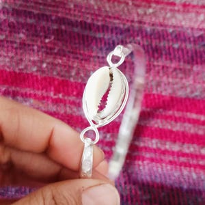 Może przedstawiać: Srebrna bransoletka z naturalną muszlą jako centralnym elementem. Bransoletka ma prosty, elegancki design z polerowanym wykończeniem. Muszla jest biała i teksturowana, dodając biżuterii odrobinę nadmorskiego uroku.