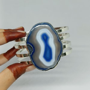 May include: A cuff bracelet featuring a large, oval-shaped blue and gray agate slice as the centerpiece. The stone displays concentric bands of color and is mounted on a silver-toned metal band composed of four parallel, thin, curved metal strips.