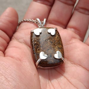 Peut inclure: Un collier pendentif rectangulaire avec une pierre brune et des accents argentés. Les accents argentés comprennent quatre motifs en forme de cœur. Le pendentif est suspendu à une chaîne argentée.