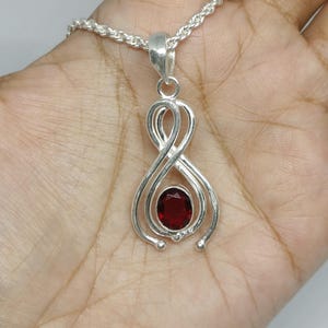 May include: A silver pendant necklace featuring a deep red oval-cut gemstone set within a polished silver frame that forms an infinity-like loop design. The pendant hangs from a silver rope chain, showcasing a metallic luster and smooth finish.