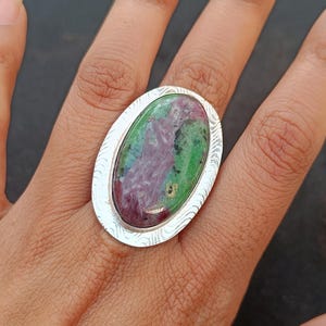 Puede incluir: Anillo ovalado con una piedra preciosa grande que muestra colores verdes, rojos y negros. La piedra está engastada en una banda de metal plateado con un diseño grabado. El anillo se lleva en un dedo. La imagen está marcada con "Shot on OnePlus".