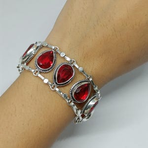 Puede incluir: Una pulsera de eslabones en tono plateado con múltiples piedras rojas facetadas en forma de lágrima, engastadas en marcos de metal trenzado. La pulsera consta de dos cadenas paralelas unidas por los engastes, creando un aspecto ancho y superpuesto.