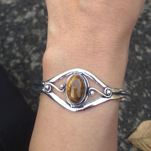 Pode incluir: Pulseira de punho prateada com uma pedra olho de tigre oval no centro. A pulseira apresenta um design vazado decorativo ao redor da pedra e é usada no pulso. A pedra é de cor castanho quente.