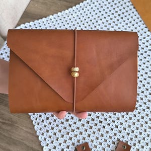May include: A tan leather envelope-style clutch with a triangular flap closure. A thin vertical leather cord wraps around the centre, secured by two small, textured, metallic gold-toned beads that act as a decorative fastener for the pouch.