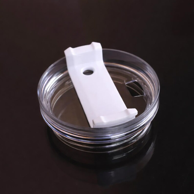 May include: A clear plastic tumbler lid with a white sliding closure. The lid has a circular design with a white plastic slider that opens and closes a drinking hole. The lid is designed to fit on a tumbler.