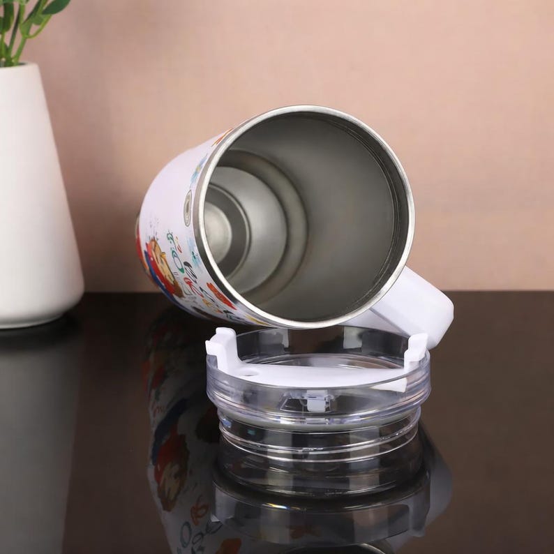 May include: White insulated travel mug with a colorful cartoon design. The mug has a white handle and a clear plastic lid. The interior is stainless steel. The mug is designed to keep beverages hot or cold.