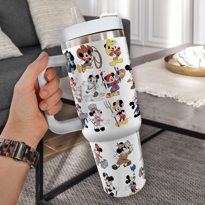 May include: White insulated tumbler featuring various Mickey Mouse character designs in different outfits. The tumbler has a white handle, lid, and straw. The tumbler is designed to keep beverages cold or hot.