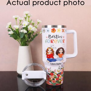May include: White travel mug with a handle, lid, and floral design. The mug features the text "Besties Forever" and illustrations of two women. The bottom of the mug has a colorful floral pattern.