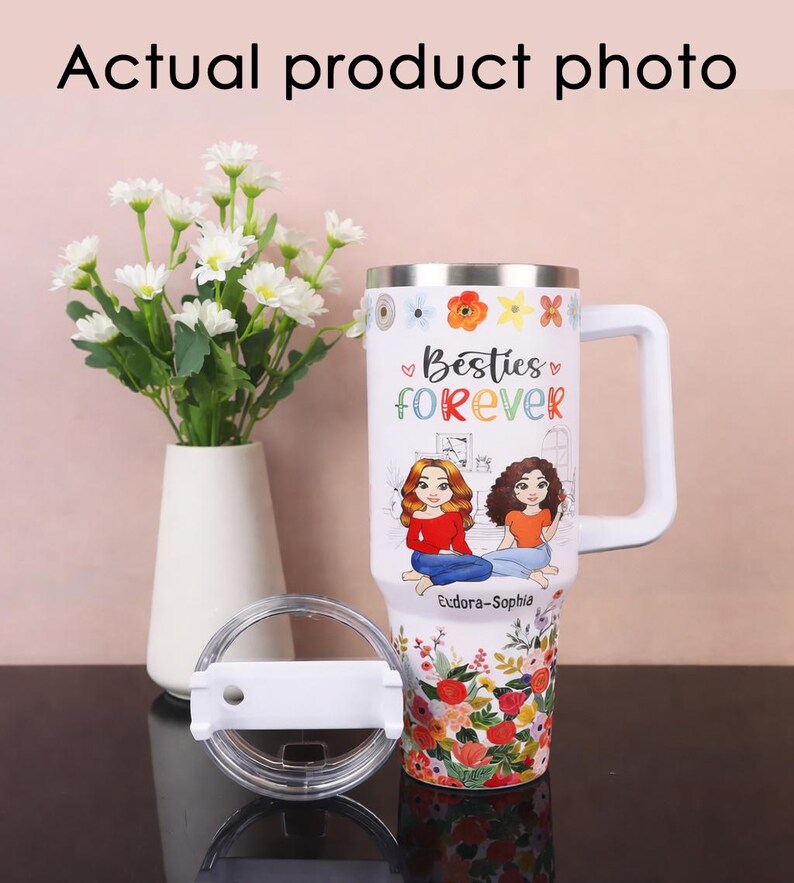 May include: White travel mug with a handle, lid, and floral design. The mug features the text "Besties Forever" and illustrations of two women. The bottom of the mug has a colorful floral pattern.