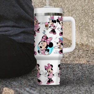 May include: White travel mug featuring a repeating pattern of Minnie Mouse in various poses, wearing pink dresses and bows. The mug has a white handle, lid, and straw. The background is a neutral color.