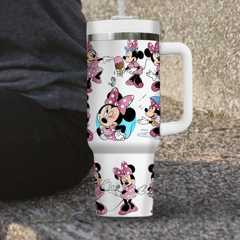 May include: White travel mug featuring a repeating pattern of Minnie Mouse in various poses, wearing pink dresses and bows. The mug has a white handle, lid, and straw. The background is a neutral color.