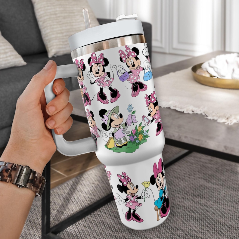 May include: White insulated tumbler with a handle and straw, featuring a repeating pattern of Minnie Mouse in various poses. Minnie is depicted in pink dresses and bows, with black accents. The tumbler has a white lid and handle.