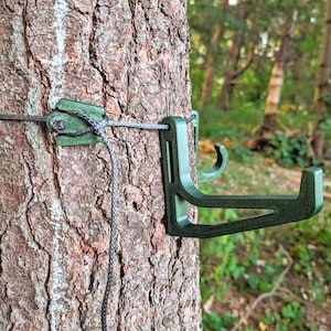 May include: A green tree stand attached to a tree trunk with a rope. The stand has a hook shape and is designed to hold items. The tree's bark is brown and textured, with a forest background.