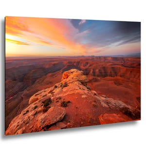May include: A rectangular wall art print featuring a high-angle landscape view of a desert canyon at sunset. The foreground shows rocky terrain in shades of orange and red, while the sky displays vibrant streaks of yellow, orange, and blue clouds.