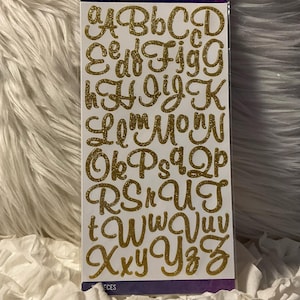 Puede incluir: Un paquete de pegatinas de alfabeto con purpurina dorada. Las pegatinas están en una fuente cursiva e incluyen las letras de la A a la Z. El paquete tiene la marca "sticko" y la palabra "STICKERS" impresa en la parte superior.